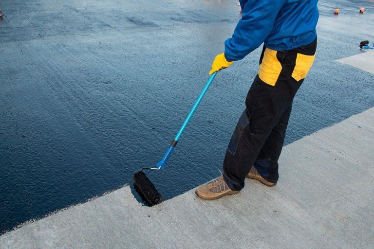 Worker rolling black waterproofing coating across a concrete deck with a roller