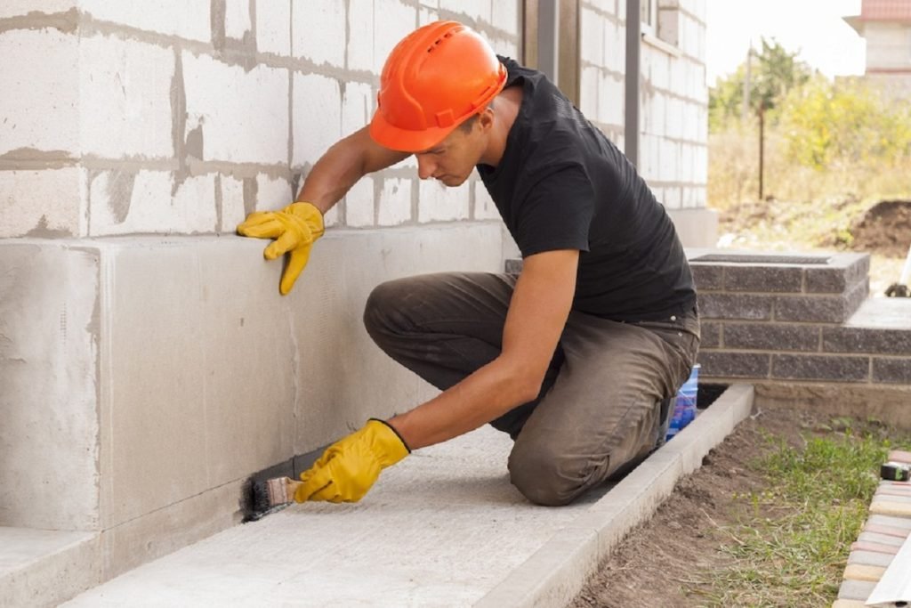 Worker in hard hat brushing sealant along an exterior foundation wall