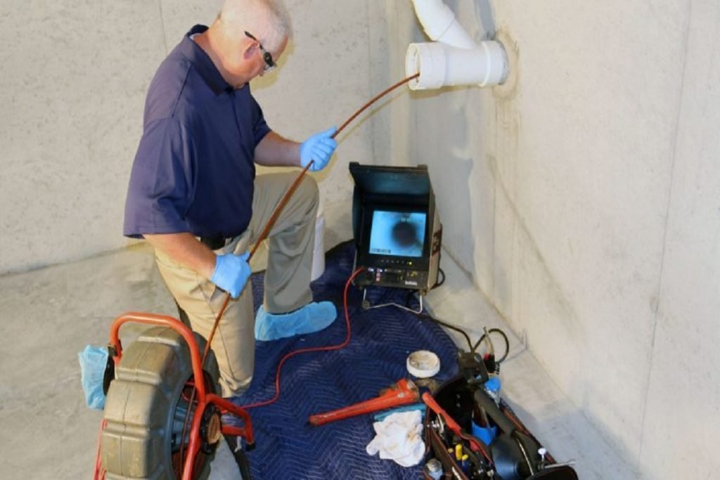Plumber feeding an inspection camera cable into a wall pipe beside a monitor