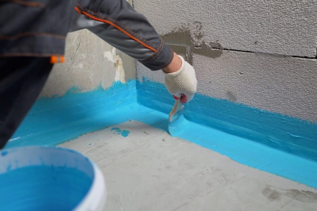Gloved hand brushing blue waterproofing membrane along a basement floor-to-wall joint