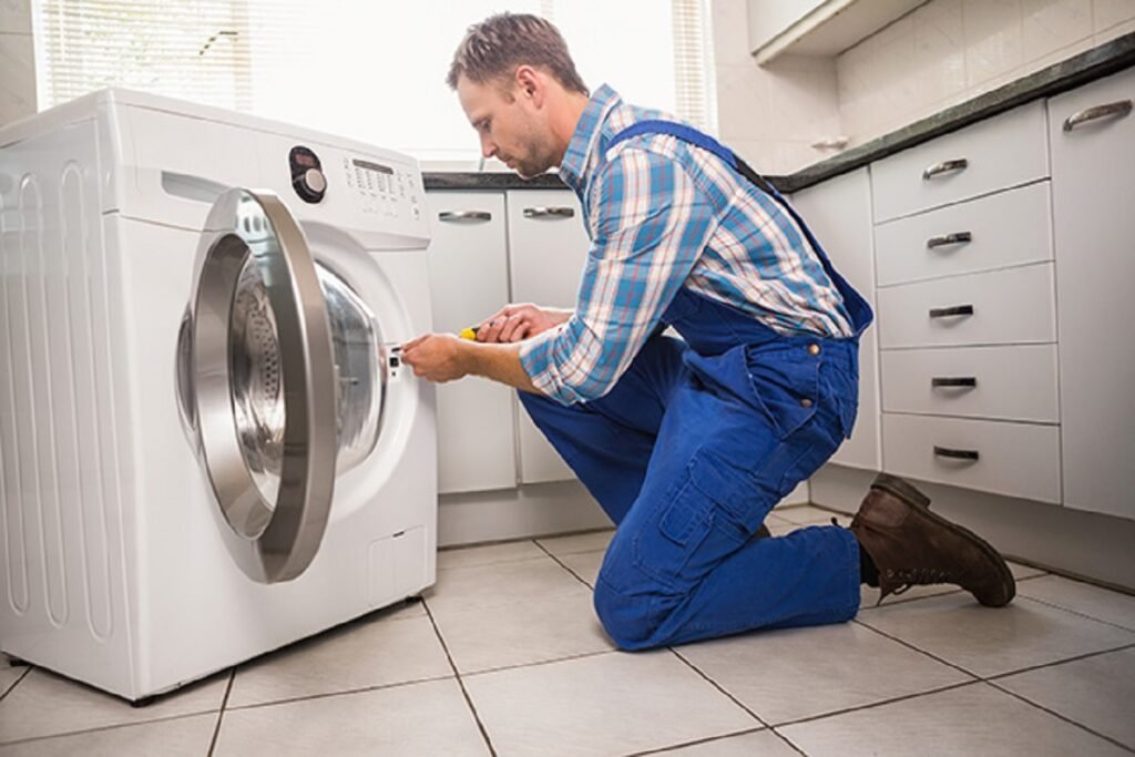Technician in blue overalls kneeling to repair a front-loading washing machine