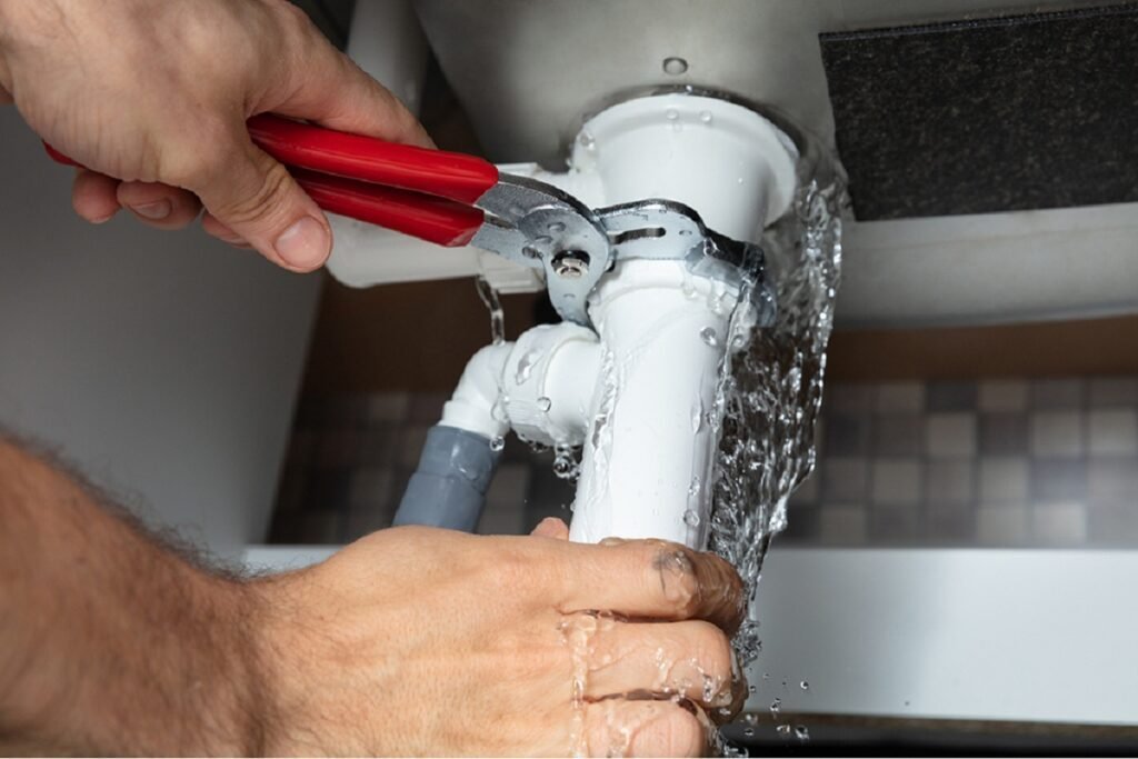 Hands using pliers on a leaking white sink drain trap spraying water