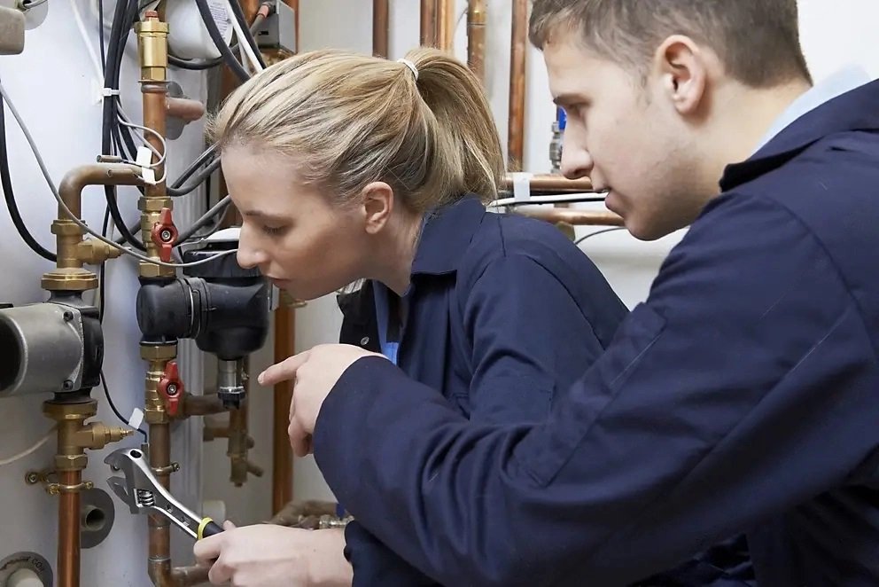 Two technicians in navy overalls inspecting copper pipework on a central heating boiler