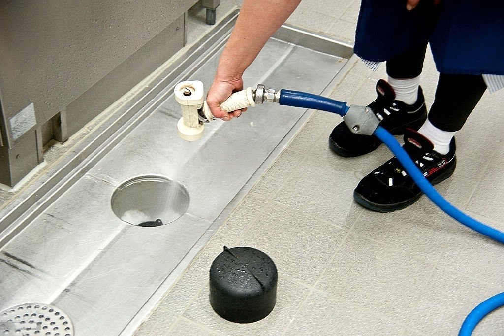 Worker rinsing a stainless steel floor drain with a spray nozzle and hose