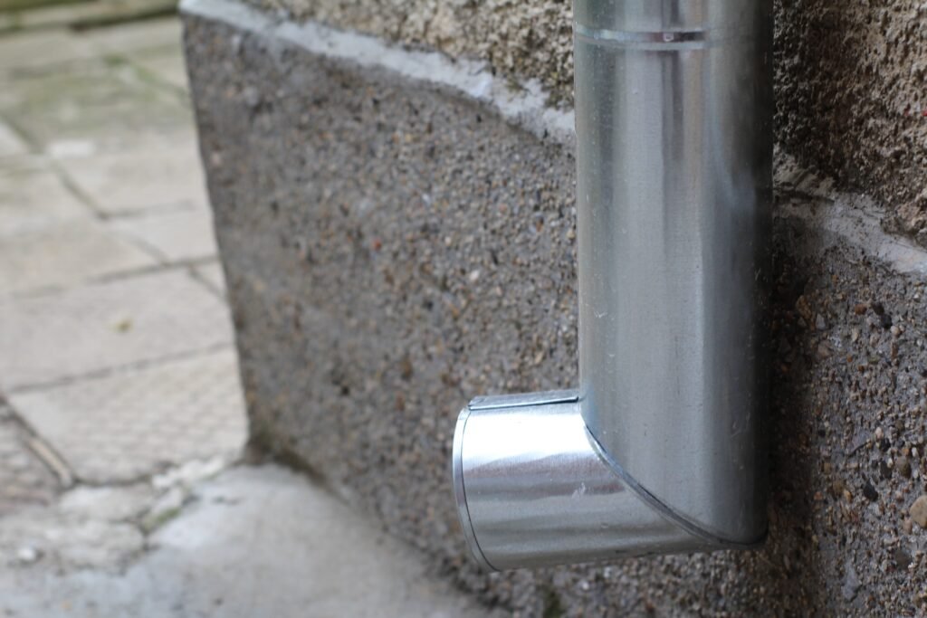 Galvanized metal downspout elbow discharging at the base of a concrete wall